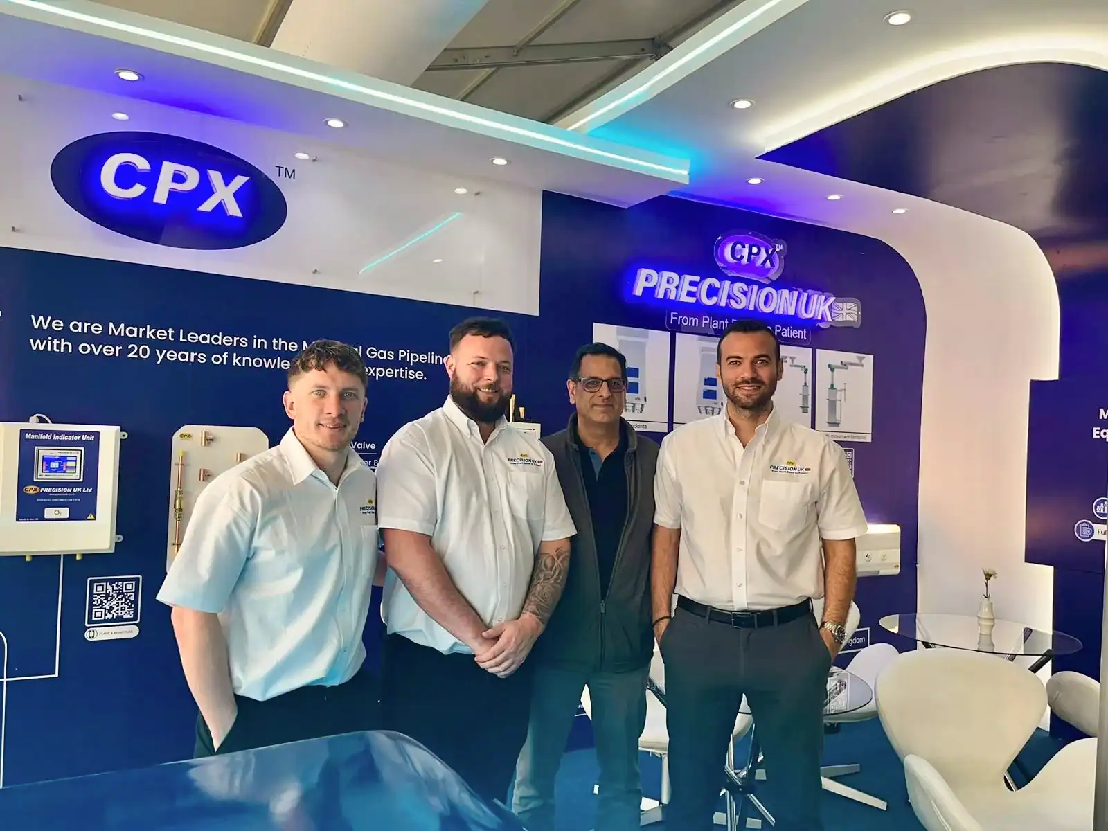 4 men (3 Precision UK colleagues and one visitor) stood in front of a white and navy exhibition stand with mounted MGPS equipment on the wall.