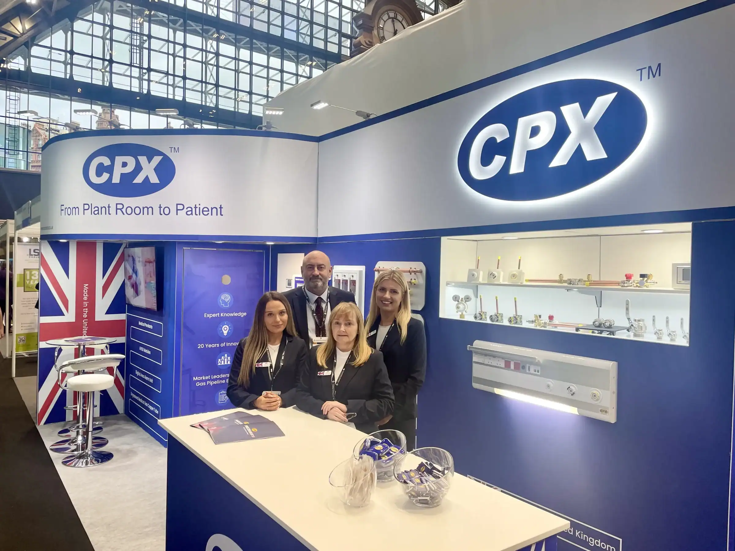 Four team members from Precision UK in navy suits stood behind desk on a blue and white exhibition stand in Manchester Central.